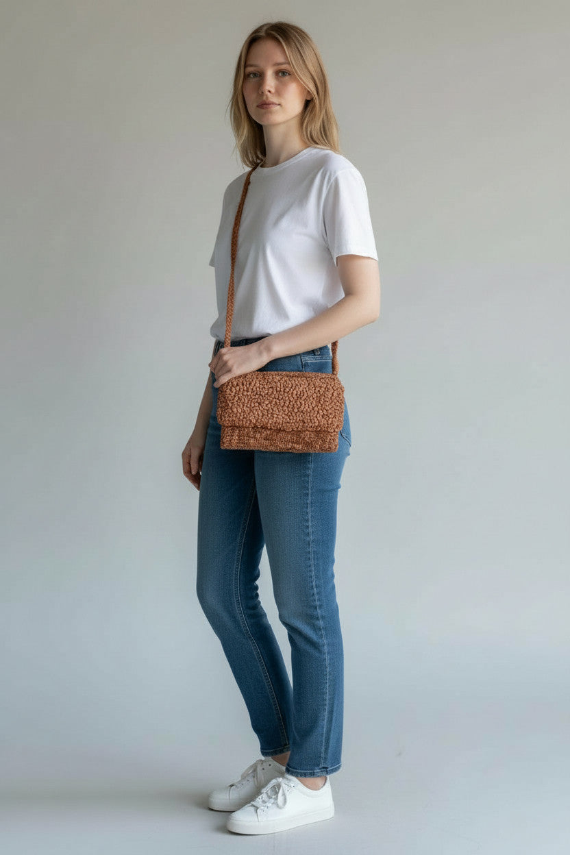 Woman wearing a white t-shirt, blue jeans, and white sneakers, holding a brown woven handbag against a plain background.